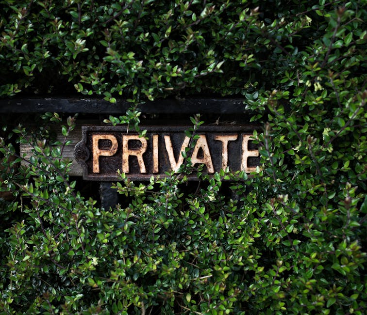 A close-up image of a weathered, rust-colored metal sign with the word 'PRIVATE' embossed on it, mounted on a wooden surface. Surrounding the sign, dense green foliage with small, glossy leaves partia