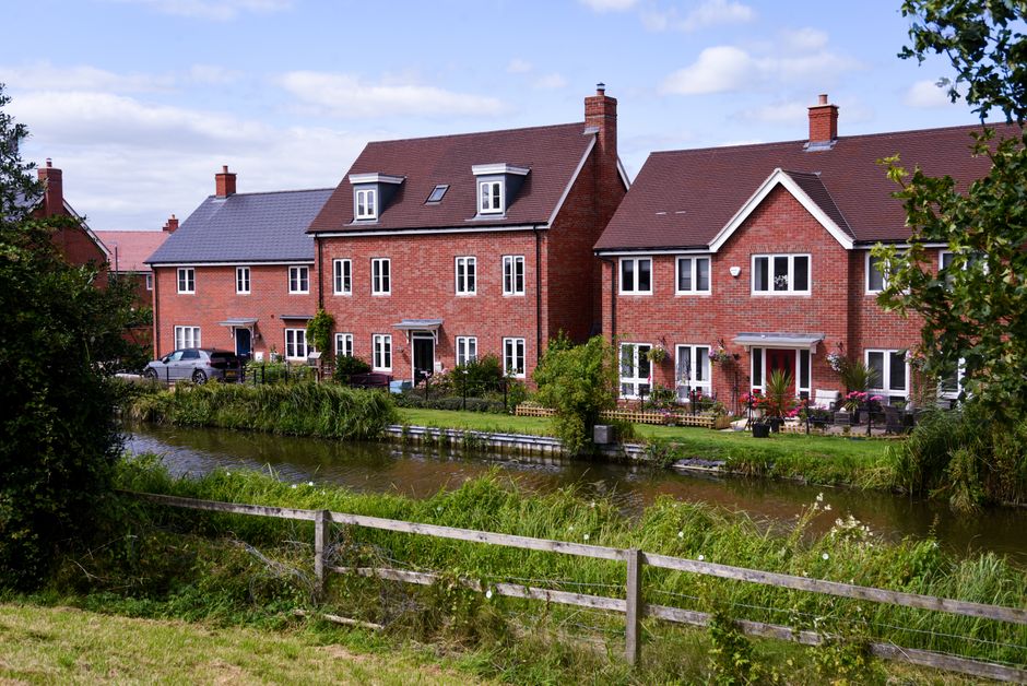 A scenic view of a residential area near a small river, featuring three red-brick houses with pitched roofs and white-framed windows. The houses are surrounded by lush, well-maintained gardens with vi